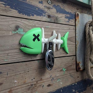 May include: A green and white fishbone-shaped key holder with a black X on the fish's face. A car key is hanging from the key holder. The key holder is mounted on a weathered wooden surface.