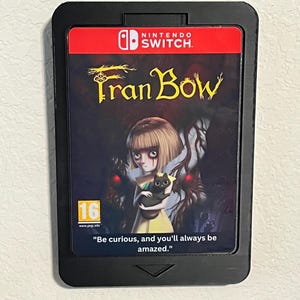 May include: A black Nintendo Switch game case with a red and white Nintendo Switch logo. The game case features a dark background with a girl holding a black cat. The game title is "Fran Bow" in yellow text. The game is rated 16+ by PEGI. The text "Be curious, and you'll always be amazed." is printed on the bottom of the case.