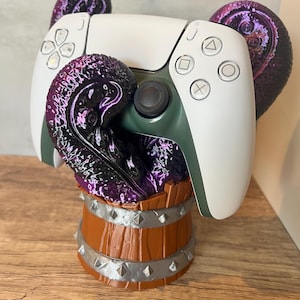 May include: A white and green PlayStation 5 controller sits on a brown wooden barrel-shaped stand, decorated with purple tentacle-like features. The stand has silver metal accents and studs. The controller's buttons are visible.