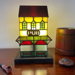 May include: A stained glass lamp shaped like a pub building with a lit interior. The lamp is on a wooden table with a wooden beer stein and a pipe.