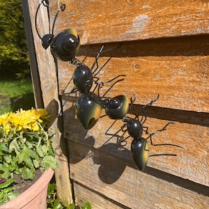 May include: Three black metal ants with orange accents are climbing up a wooden wall. The ants are made of wire and have a realistic appearance.