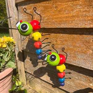 May include: Two colourful metal caterpillars with googly eyes and black wire legs. The caterpillars are attached to a wooden surface.