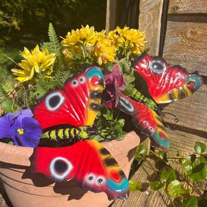 May include: Two red, yellow, black and blue metal butterflies with black eyes and a green and yellow body. The butterflies are attached to a brown flower pot with yellow flowers and green leaves.