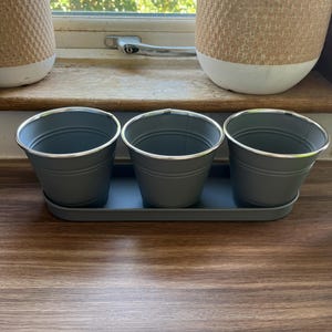 May include: Three grey metal buckets with silver rims on a matching tray. The buckets are arranged in a row, ideal for small plants or decorative items. The background includes a wooden surface and a window.