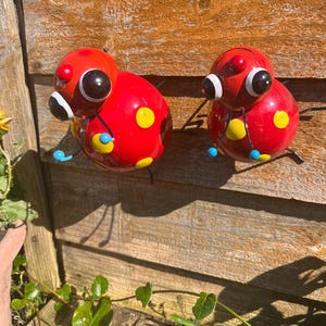 May include: Two red ladybug-shaped metal ornaments with black eyes, yellow and blue accents, and white spots. The ornaments are attached to a wooden surface.