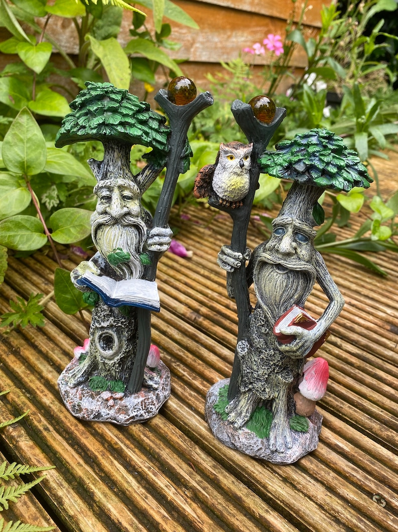 Pu&ograve; includere: Due statue da giardino fantasiose raffiguranti figure simili ad alberi con lunghe barbe e che tengono bastoni. Una figura tiene in mano un libro, l'altra ha un gufo appollaiato sul suo bastone. Le statue hanno cime verdi a forma di albero e sono su una superficie di legno.