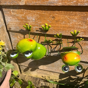 May include: Two green and yellow metal frogs with large eyes hanging on a wooden fence. The frogs have a textured finish and are in a playful pose.