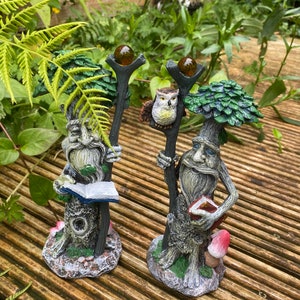 Pu&ograve; includere: Due statue da giardino fantasiose di figure simili ad alberi con lunghe barbe e che tengono bastoni sormontati da globi color ambra. Una figura tiene un libro, l'altra ha un gufo. Le statue sono su un ponte di legno.