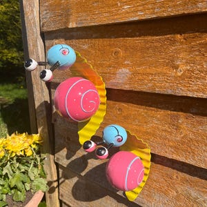 May include: Three metal snails with yellow, pink and blue paint. The snails are attached to a wooden surface.