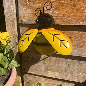 May include: A yellow and black metal bee garden decoration with wire antennae. The bee is attached to a wooden fence.