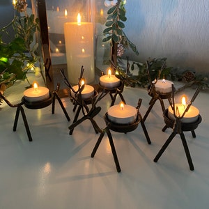 May include: Six black metal reindeer candle holders with white tea lights. The candle holders are arranged in a circle on a white surface.