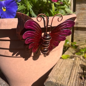 May include: A pink metal butterfly garden decoration with copper accents. The butterfly is attached to a terracotta flower pot.