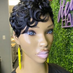 May include: A mannequin head with a short, curly black wig. The mannequin has blue eyes and is wearing long, bright yellow earrings. The background includes a green wall and a sign that says "HAIR".