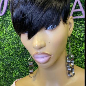 May include: A mannequin head with short, black hair styled in a side-swept bang. The mannequin has blue eyes, full lips, and is wearing silver earrings. The background is a green wall with a purple logo.