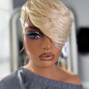 May include: A blonde, short-layered wig with side-swept bangs. The wig is styled on a mannequin head with detailed makeup, including dark eyeshadow and full eyelashes. The mannequin is wearing a gray off-the-shoulder top.