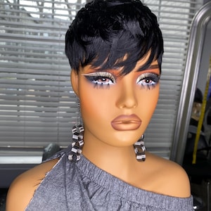 May include: A mannequin head with a short, black, wavy wig. The mannequin has dramatic eye makeup with white eyeliner and silver eyeshadow. It is wearing silver and clear geometric earrings and a gray off-the-shoulder top.