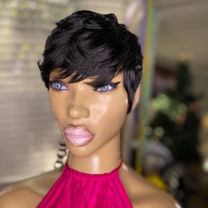 May include: A mannequin head with a short, dark, layered hairstyle. The mannequin has blue eyes, full lips, and is wearing a bright pink halter top. The background is blurred, with a glimpse of a window and other objects.