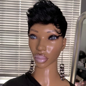 Puede incluir: Una cabeza de maniquí con cabello negro corto peinado en un corte pixie. El maniquí tiene piel clara, ojos azules y labios carnosos. Lleva pendientes largos y colgantes con un diseño a rayas en blanco y negro. El maniquí lleva una camiseta negra.