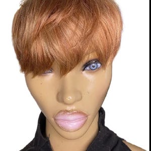 Puede incluir: Cabeza de maniquí con una peluca corta y cobriza. El maniquí tiene la piel clara, ojos azules y labios rosados. Lleva un top negro. El fondo es blanco.