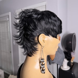 Puede incluir: Una cabeza de maniquí con un peinado corto y negro con puntas y pendientes de cadena plateada. Los pendientes tienen una serie de piezas metálicas curvas y conectadas. El maniquí está en un salón.