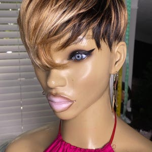 May include: A mannequin head displaying a short, layered wig with a blend of brown, blonde, and black tones. The mannequin wears a bright pink halter top and gold earrings. The wig features a side-swept bang.