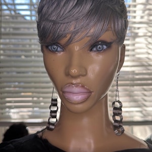 Puede incluir: Una cabeza de maniquí con una peluca corta gris y ojos azules. El maniquí lleva pendientes de cadena plateada y una parte superior negra. El fondo incluye una ventana con persianas.