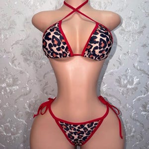 May include: A two-piece bikini set with a leopard print pattern. The bikini top has a halter neck design with red trim, and the bottoms are a classic thong style with matching red ties. The swimsuit is displayed on a mannequin.