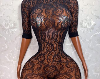 May include: Black lace bodysuit with short sleeves and shorts. The bodysuit features a floral pattern and is displayed on a mannequin. The mannequin has a brown skin tone and is wearing iridescent pasties.