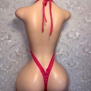 May include: A vibrant pink bodysuit featuring a halter neck design and a thong back. The edges and straps are embellished with small, gold-toned studs. The garment is displayed on a mannequin.