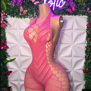 May include: A vibrant pink halter-neck bodysuit with a distinctive open-weave pattern. The garment showcases a geometric design and cut-out features, creating a striking visual effect. The word "Votic" is visible in neon lights in the background.