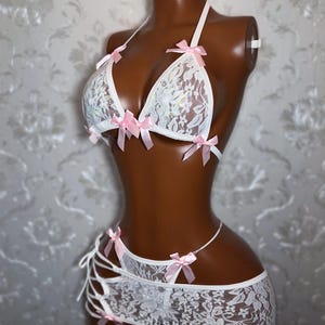 Peut inclure: Ensemble de lingerie en dentelle blanche avec des n&oelig;uds en satin rose. L'ensemble comprend un soutien-gorge triangle, une jupe et un string. Le soutien-gorge et la jupe sont orn&eacute;s de n&oelig;uds roses d&eacute;licats. La lingerie est pr&eacute;sent&eacute;e sur un mannequin.