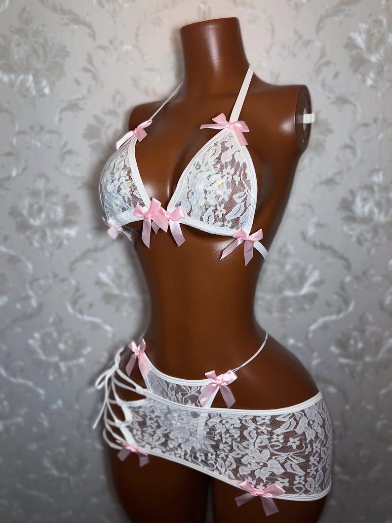 Peut inclure: Ensemble de lingerie en dentelle blanche avec des n&oelig;uds en satin rose. L'ensemble comprend un soutien-gorge triangle, une jupe et un string. Le soutien-gorge et la jupe sont orn&eacute;s de n&oelig;uds roses d&eacute;licats. La lingerie est pr&eacute;sent&eacute;e sur un mannequin.