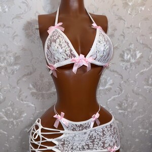 Peut inclure: Ensemble de lingerie en dentelle blanche avec des n&oelig;uds roses. L'ensemble comprend un haut dos nu, une jupe &agrave; lacets sur les c&ocirc;t&eacute;s et un string assorti. La lingerie est pr&eacute;sent&eacute;e sur un mannequin.