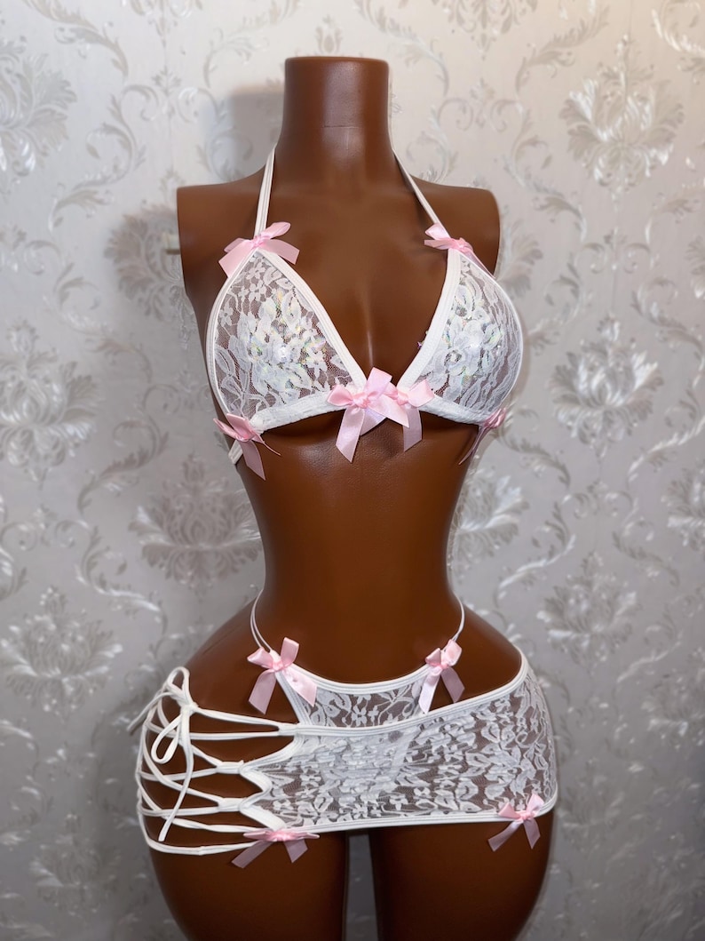 Peut inclure: Ensemble de lingerie en dentelle blanche avec des n&oelig;uds roses. L'ensemble comprend un haut dos nu, une jupe &agrave; lacets sur les c&ocirc;t&eacute;s et un string assorti. La lingerie est pr&eacute;sent&eacute;e sur un mannequin.