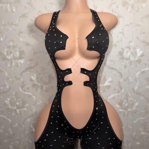 May include: Black bodysuit with rhinestone embellishments. The halter-style top features cut-out details and a criss-cross design. The shorts-style bottom has cut-out sides. The bodysuit is displayed on a mannequin.