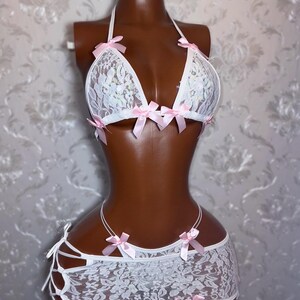 Peut inclure: Ensemble de lingerie en dentelle blanche avec des n&oelig;uds en satin rose. L'ensemble comprend un soutien-gorge &agrave; licou et une mini-jupe avec des d&eacute;tails de liens. Le soutien-gorge et la jupe sont orn&eacute;s de d&eacute;licats n&oelig;uds roses. La lingerie est pr&eacute;sent&eacute;e sur un mannequin.