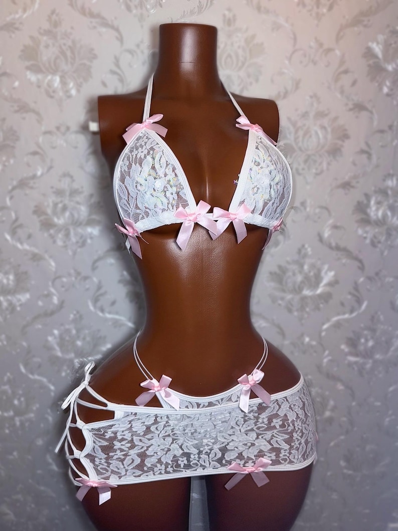 Peut inclure: Ensemble de lingerie en dentelle blanche avec des n&oelig;uds en satin rose. L'ensemble comprend un soutien-gorge &agrave; licou et une mini-jupe avec des d&eacute;tails de liens. Le soutien-gorge et la jupe sont orn&eacute;s de d&eacute;licats n&oelig;uds roses. La lingerie est pr&eacute;sent&eacute;e sur un mannequin.