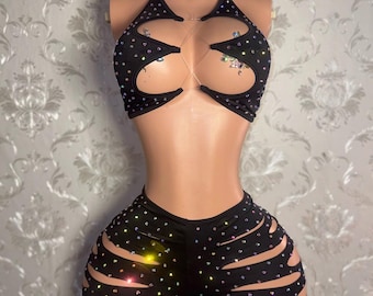 May include: A black two-piece set consisting of a halter-style crop top with circular cutouts and matching shorts with horizontal side slits. The entire garment is embellished with small, multicolored iridescent rhinestones that reflect light.