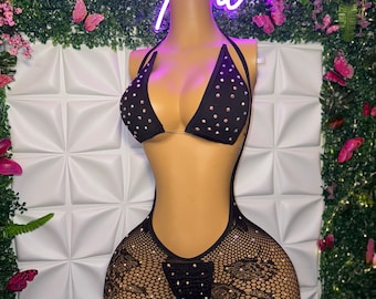 May include: Black swimsuit with rhinestone detailing. The top is a triangle bikini, and the bottom is a one-piece with a fishnet design. The swimsuit is displayed on a mannequin against a white textured wall.