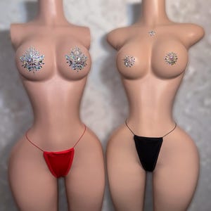 Op de afbeelding: Twee mannequin torso's met strass tepel pasties en string ondergoed. Eén mannequin draagt een rode string, de andere een zwarte string. De mannequins hebben een lichte huidskleur tegen een neutrale achtergrond.