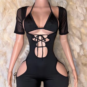 May include: Black halter-neck bodysuit with a cutout design and lace-up detail. The bodysuit features a triangle top and short sleeves. The shorts have side cutouts with tie closures. The outfit is displayed on a mannequin.