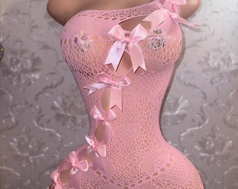 May include: Pink one-shoulder bodysuit with a fishnet design. The bodysuit features multiple pink satin bows and rhinestone embellishments. The garment has a shorts-style bottom and is displayed on a mannequin.
