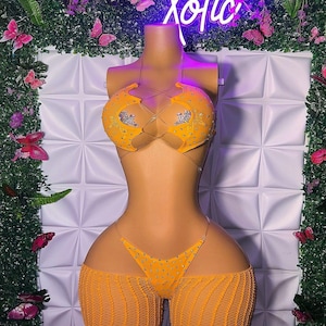 May include: A bright orange two-piece bikini set featuring a halter-style top with clear straps, intricate lace-up detailing, and silver rhinestone embellishments. The matching bottom includes a thong-style cut with rhinestones and a sheer mesh overlay.