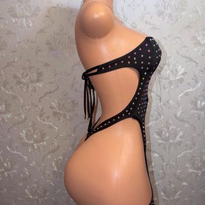 May include: Black bodysuit adorned with rhinestones. The strapless one-piece garment has cut-out sections and attached garter straps. The bodysuit is displayed on a mannequin.