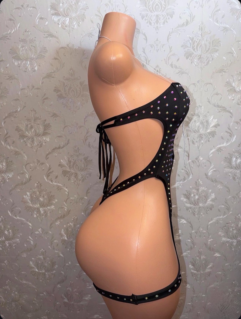 May include: Black bodysuit adorned with rhinestones. The strapless one-piece garment has cut-out sections and attached garter straps. The bodysuit is displayed on a mannequin.