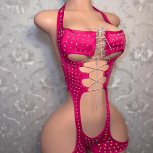 May include: A vibrant pink bodysuit with a halter neck and cut-out design. The garment is adorned with small, sparkling rhinestones and features a lace-up detail at the bust. The shorts section has cut-out sides.