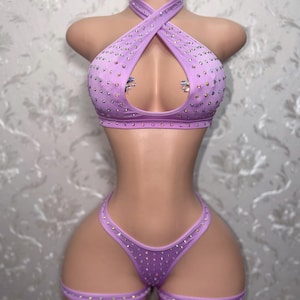 May include: A lavender two-piece set featuring a halter-style top with a crisscross neckline and a cutout center, paired with matching high-cut bottoms. Both pieces are embellished with scattered clear rhinestones and include leg garter straps.