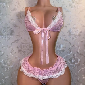 May include: Pink lace lingerie set with white trim, displayed on a mannequin. The set includes a bra with a bow and jeweled accents, a matching skirt, and panties. The background is a floral wallpaper.