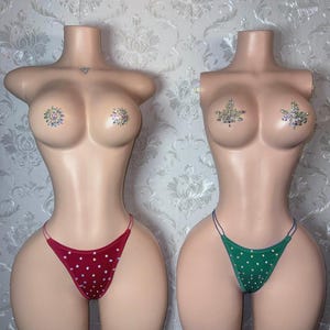 Puede incluir: Dos torsos de maniquíes sin cabeza. Un maniquí lleva un tanga rojo con lunares blancos, el otro un tanga verde con lunares blancos. Ambos maniquíes tienen pezoneras de pedrería.