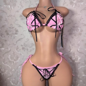 Può includere: Un set bikini a due pezzi in rosa chiaro con bordi in pizzo nero e dettagli a nastro nero incrociato. Il top a triangolo e lo slip abbinato sono decorati con piccoli fiocchi rosa e sottili laccetti in nastro nero sui lati dello slip.